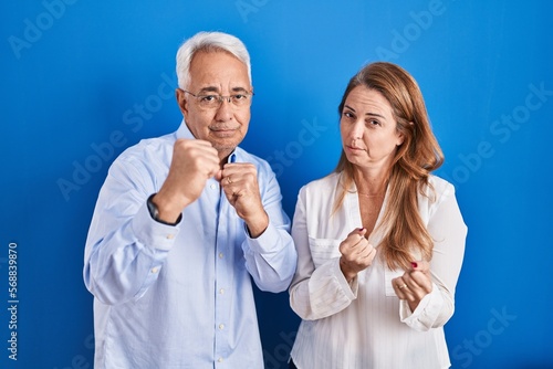 Middle age hispanic couple standing over blue background ready to fight with fist defense gesture, angry and upset face, afraid of problem