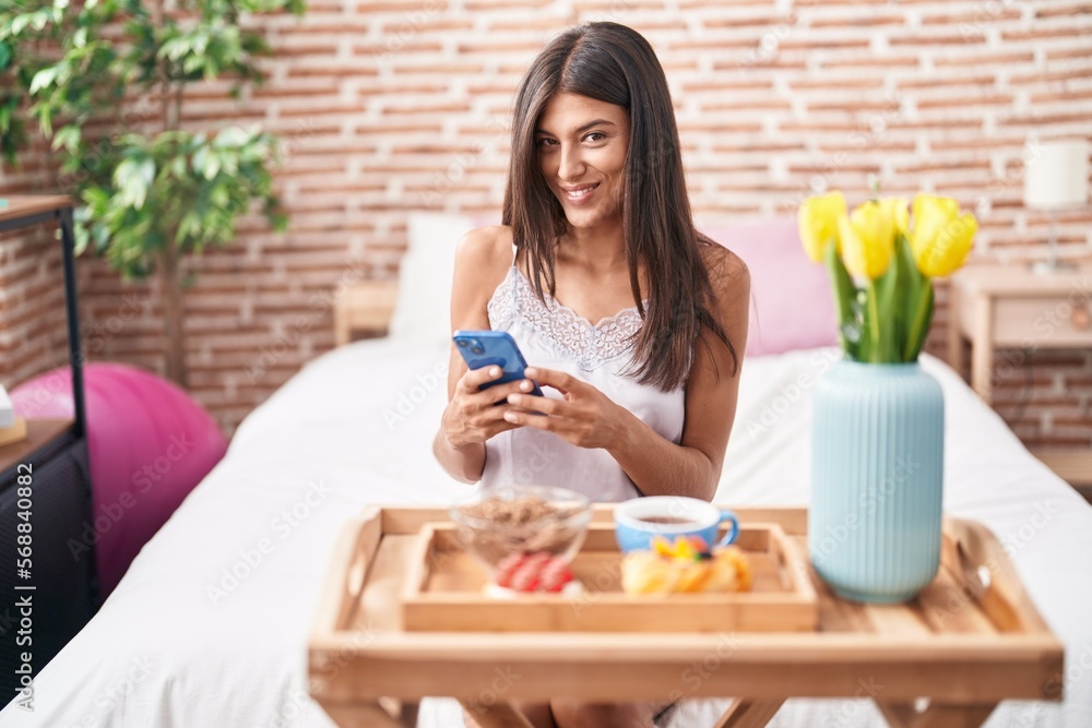 Young beautiful hispanic woman having gift breakfast using smartphone at bedroom