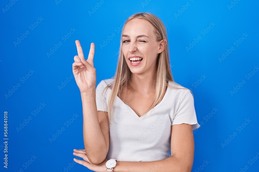 Fototapeta premium Young caucasian woman standing over blue background smiling with happy face winking at the camera doing victory sign with fingers. number two.