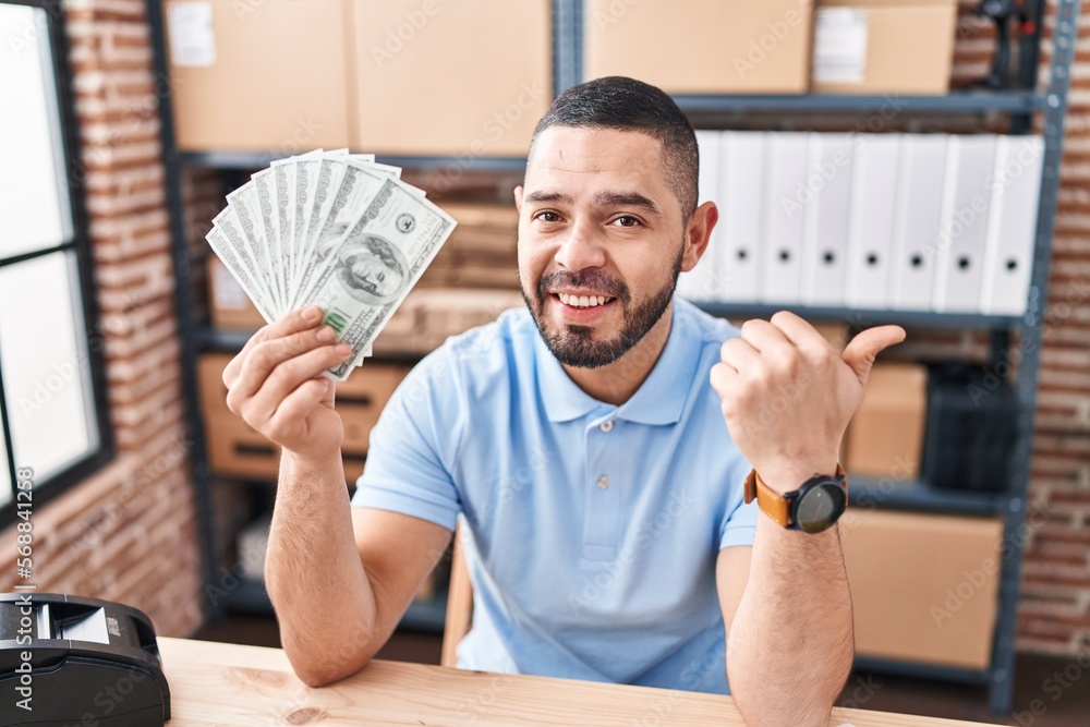 Hispanic man working at small business ecommerce holding dollars pointing thumb up to the side smiling happy with open mouth Hispanic man working at small business ecommerce holding dollars pointing thumb up to the side smiling happy with open mouth