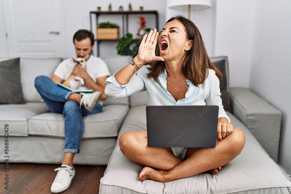 Hispanic middle age couple at home, woman using laptop shouting and screaming loud to side with hand on mouth. communication concept. Hispanic middle age couple at home, woman using laptop shouting and screaming loud to side with hand on mouth. communication concept.