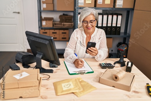 Middle age woman ecommerce business worker using smartphone write on clipboard at office