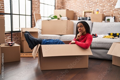 Young african american with braids moving to a new home inside of a cardboard box serious face thinking about question with hand on chin, thoughtful about confusing idea © Krakenimages.com