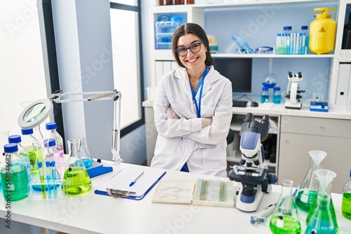 Young hispanic woman wearing scientist uniform sitting with arms crossed gesture at laboratory
