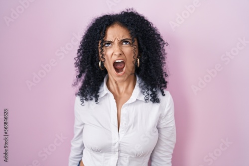 Hispanic woman with curly hair standing over pink background angry and mad screaming frustrated and furious, shouting with anger. rage and aggressive concept.