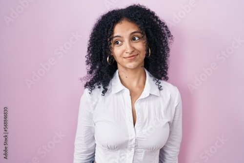 Hispanic woman with curly hair standing over pink background smiling looking to the side and staring away thinking.