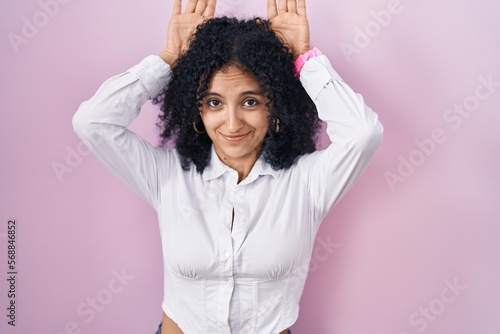 Hispanic woman with curly hair standing over pink background doing bunny ears gesture with hands palms looking cynical and skeptical. easter rabbit concept. © Krakenimages.com