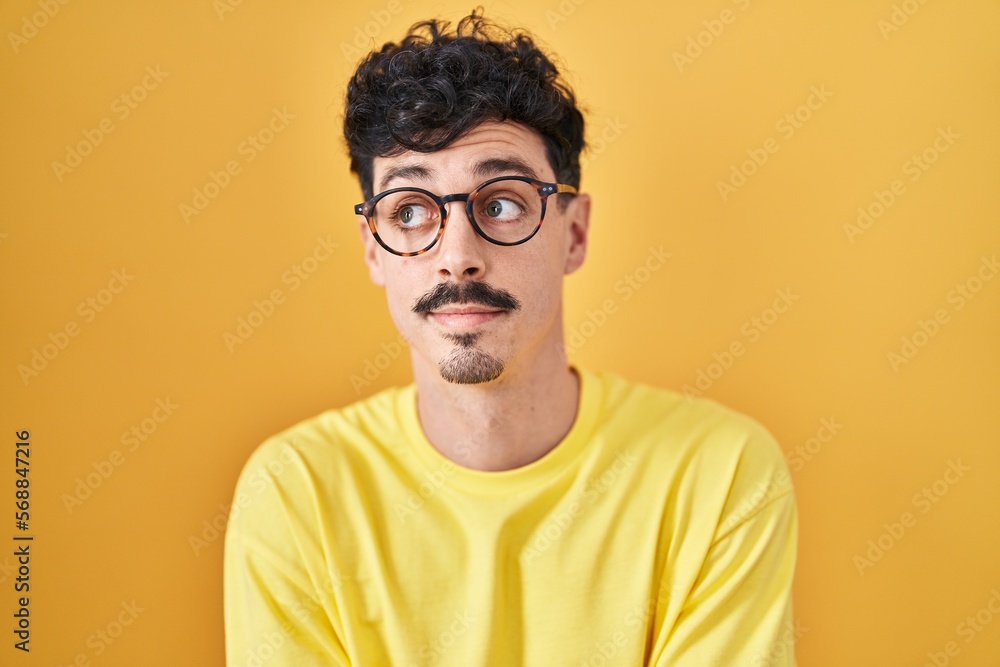 Hispanic man wearing glasses standing over yellow background smiling looking to the side and staring away thinking. Hispanic man wearing glasses standing over yellow background smiling looking to the side and staring away thinking.