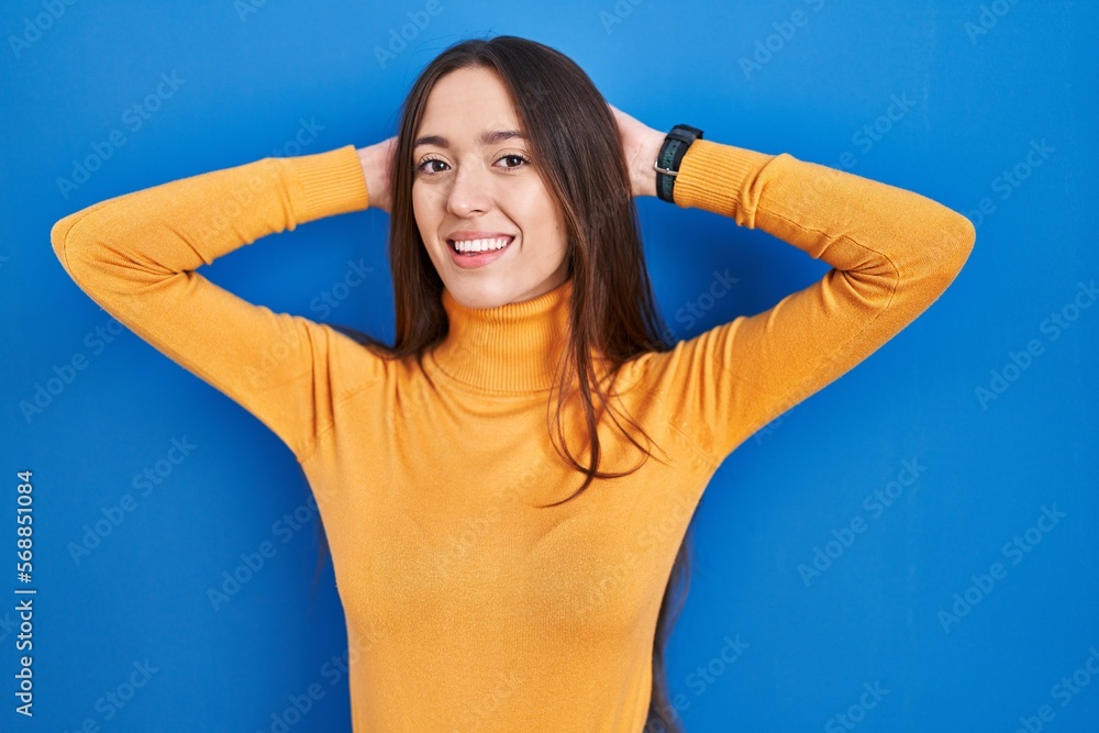 Young brunette woman standing over blue background relaxing and stretching, arms and hands behind head and neck smiling happy Young brunette woman standing over blue background relaxing and stretching, arms and hands behind head and neck smiling happy