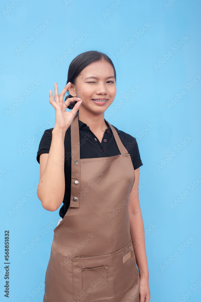 A young asian female waitress or barista winks while making the ok sign. Wearing a brown apron and black shirt welcoming a customer and against a light blue background. A young asian female waitress or barista winks while making the ok sign. Wearing a brown apron and black shirt welcoming a customer and against a light blue background.