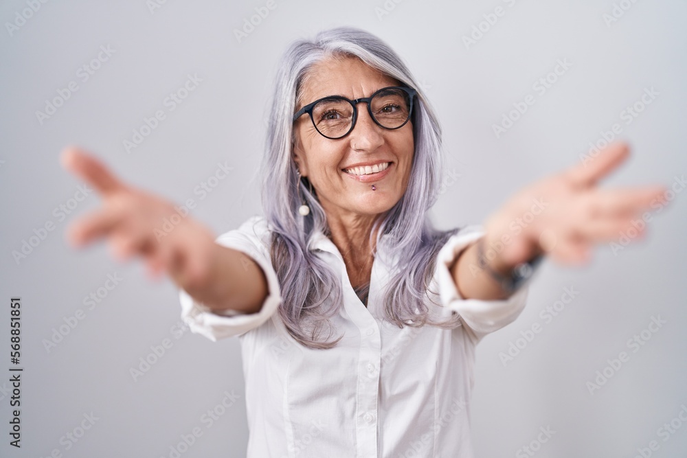 Middle age woman with tattoos wearing glasses standing over white background looking at the camera smiling with open arms for hug. cheerful expression embracing happiness. Middle age woman with tattoos wearing glasses standing over white background looking at the camera smiling with open arms for hug. cheerful expression embracing happiness.