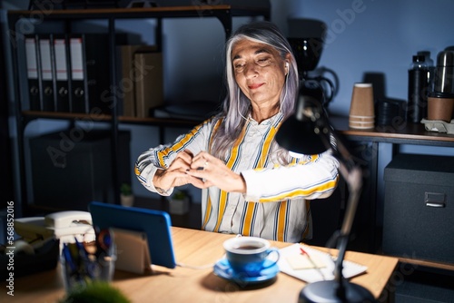 Middle age woman with grey hair working at the office at night smiling in love doing heart symbol shape with hands. romantic concept.
