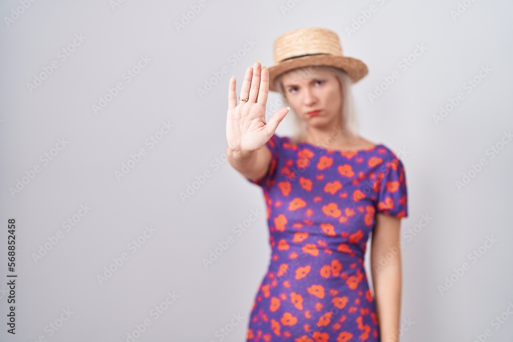 Young caucasian woman wearing flowers dress and summer hat doing stop sing with palm of the hand. warning expression with negative and serious gesture on the face. Young caucasian woman wearing flowers dress and summer hat doing stop sing with palm of the hand. warning expression with negative and serious gesture on the face.