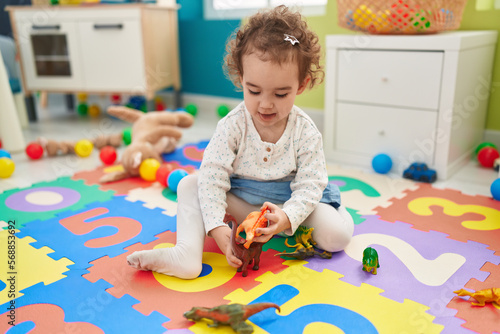 Adorable hispanic toddler playing with dino toy sitting on floor at kindergarten