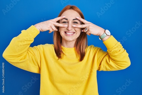 Young woman standing over blue background doing peace symbol with fingers over face, smiling cheerful showing victory © Krakenimages.com