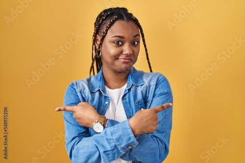 African american woman with braids standing over yellow background pointing to both sides with fingers, different direction disagree photo