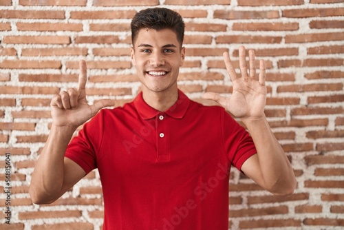 Young hispanic man standing over bricks wall showing and pointing up with fingers number seven while smiling confident and happy.