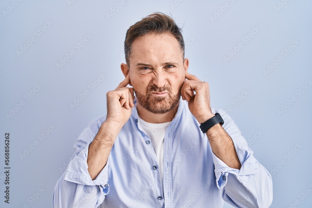Middle age caucasian man standing over blue background covering ears with fingers with annoyed expression for the noise of loud music. deaf concept. Middle age caucasian man standing over blue background covering ears with fingers with annoyed expression for the noise of loud music. deaf concept.