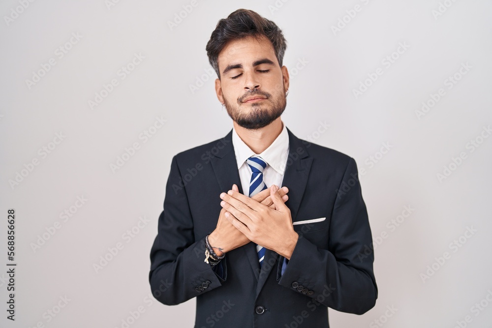 Young hispanic man with tattoos wearing business suit and tie smiling with hands on chest with closed eyes and grateful gesture on face. health concept. Young hispanic man with tattoos wearing business suit and tie smiling with hands on chest with closed eyes and grateful gesture on face. health concept.