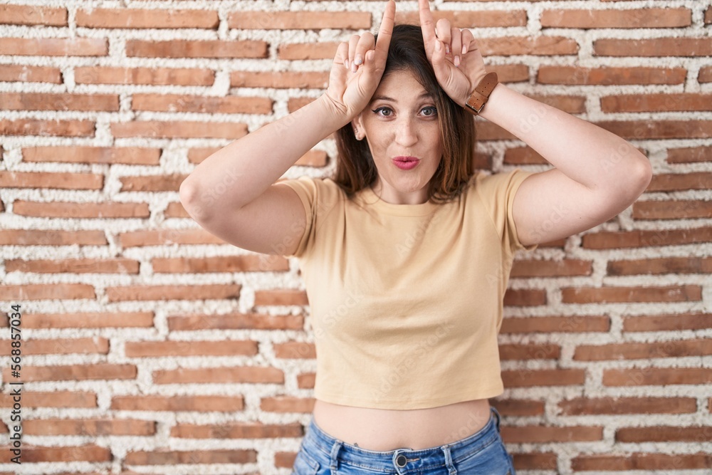 Young brunette woman standing over bricks wall doing funny gesture with finger over head as bull horns Young brunette woman standing over bricks wall doing funny gesture with finger over head as bull horns