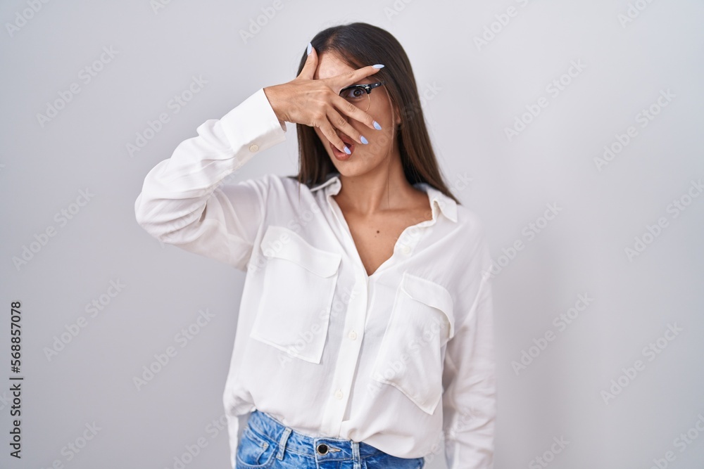 Young brunette woman wearing glasses peeking in shock covering face and eyes with hand, looking through fingers with embarrassed expression. Young brunette woman wearing glasses peeking in shock covering face and eyes with hand, looking through fingers with embarrassed expression.