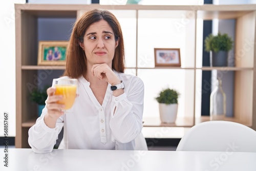 Brunette woman drinking glass of orange juice thinking worried about a question  concerned and nervous with hand on chin