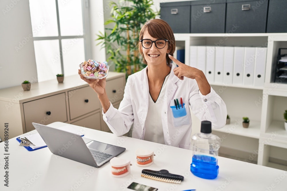 Brunette dentist woman holding sweets pointing finger to one self smiling happy and proud Brunette dentist woman holding sweets pointing finger to one self smiling happy and proud