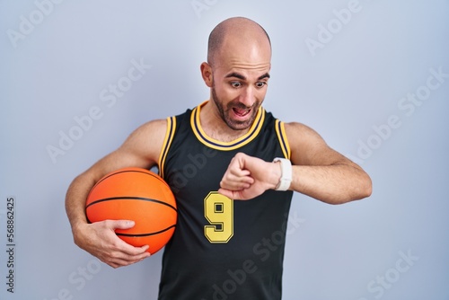 Young bald man with beard wearing basketball uniform holding ball looking at the watch time worried, afraid of getting late