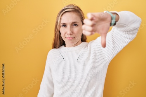 Young caucasian woman wearing white sweater over yellow background looking unhappy and angry showing rejection and negative with thumbs down gesture. bad expression.