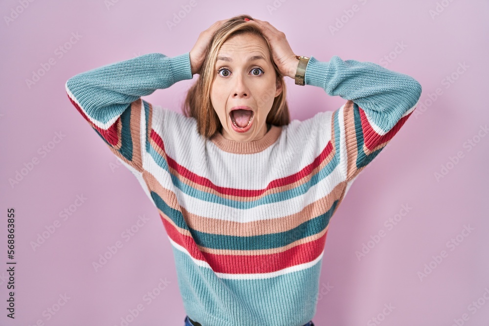 Young blonde woman standing over pink background crazy and scared with hands on head, afraid and surprised of shock with open mouth Young blonde woman standing over pink background crazy and scared with hands on head, afraid and surprised of shock with open mouth