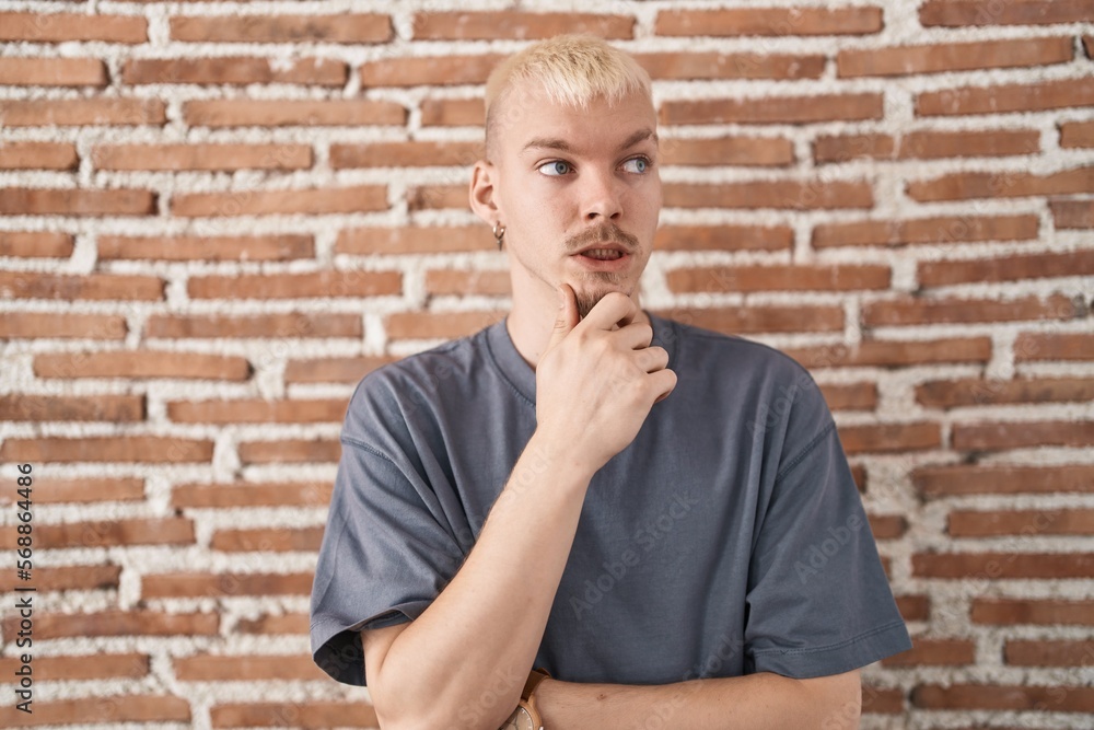 Young caucasian man standing over bricks wall thinking worried about a question, concerned and nervous with hand on chin Young caucasian man standing over bricks wall thinking worried about a question, concerned and nervous with hand on chin