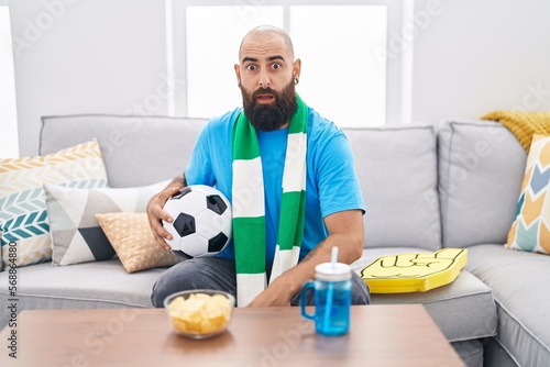 Young hispanic man with beard and tattoos football hooligan holding ball supporting team scared and amazed with open mouth for surprise, disbelief face © Krakenimages.com