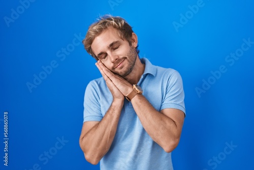 Caucasian man standing over blue background sleeping tired dreaming and posing with hands together while smiling with closed eyes.