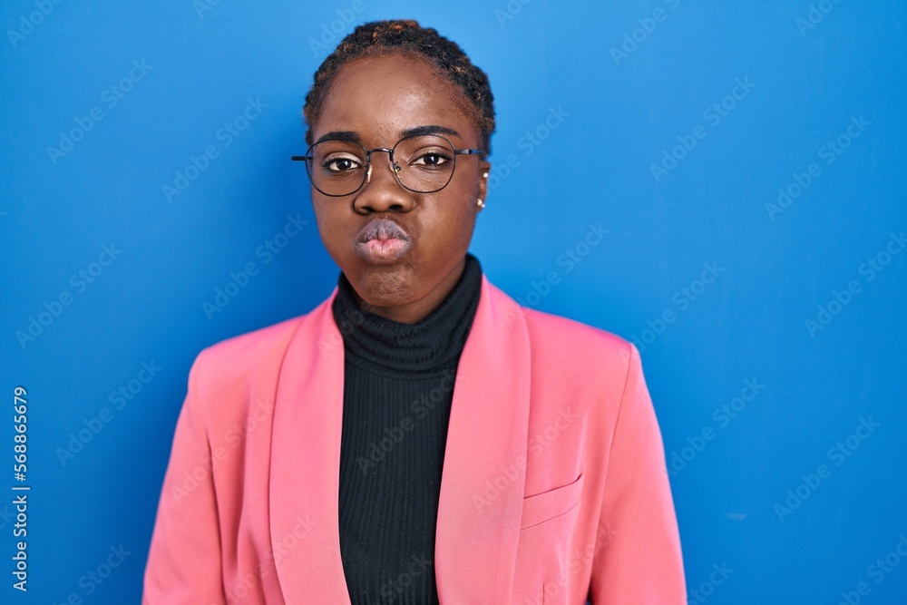 Obraz premium Beautiful black woman standing over blue background puffing cheeks with funny face. mouth inflated with air, crazy expression.