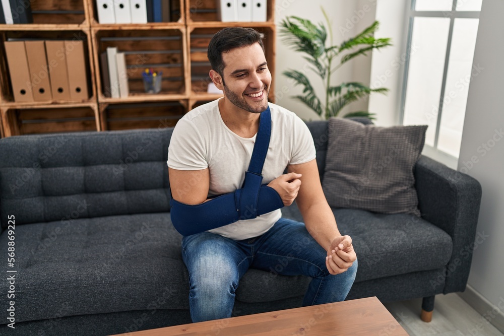 Obraz premium Young hispanic man with beard wearing arm on sling sitting at therapy consult looking positive and happy standing and smiling with a confident smile showing teeth