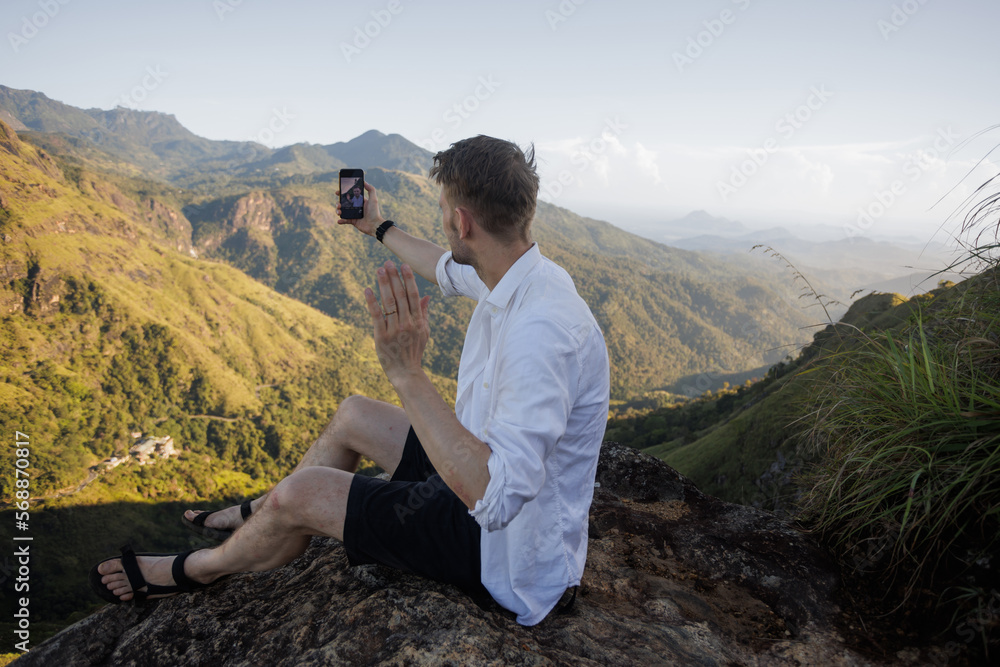 Naklejka premium Young man in nature. People and mountain. Healthy lifestyle. Happy walk in traveling in Sri Lanka. Outdoor activity casual guy with backpack. Male making travel photography with smartphone. Daily life