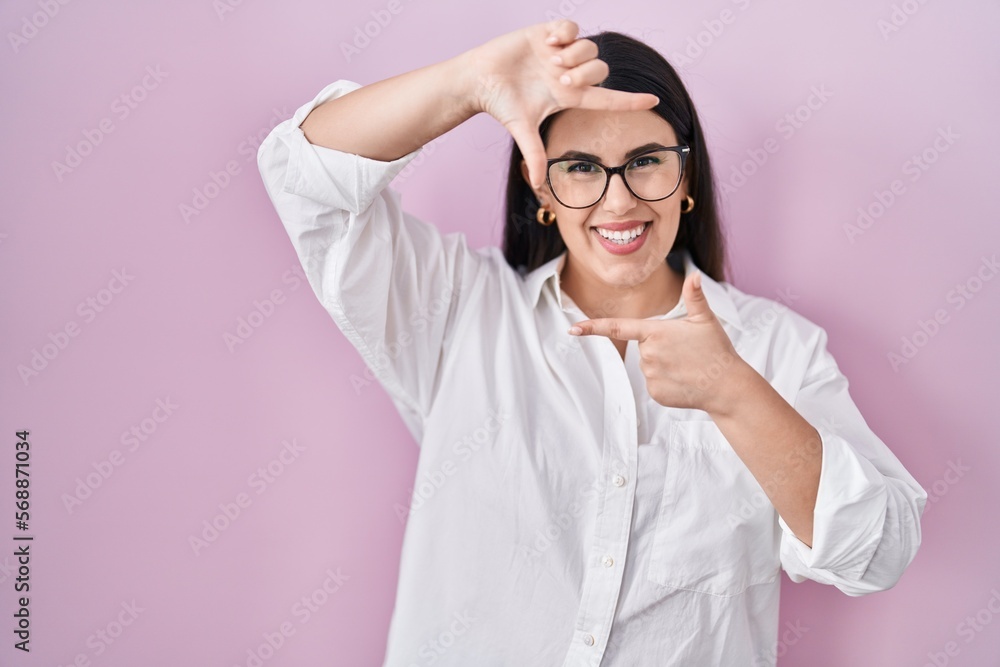 Young brunette woman standing over pink background smiling making frame with hands and fingers with happy face. creativity and photography concept.