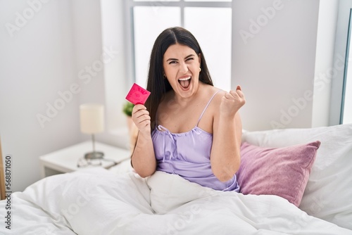 Young brunette woman wearing lingerie and holding condom on the bed screaming proud, celebrating victory and success very excited with raised arms