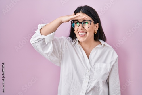Konstfotografi Young brunette woman standing over pink background very happy and smiling looking far away with hand over head