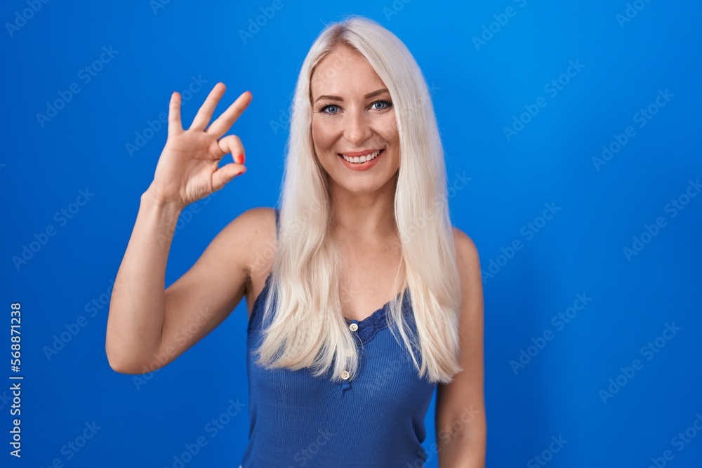Fototapeta premium Caucasian woman standing over blue background smiling positive doing ok sign with hand and fingers. successful expression.