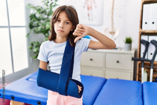 Young hispanic girl wearing arm on sling at rehabilitation clinic with angry face, negative sign showing dislike with thumbs down, rejection concept Free Download Young hispanic girl wearing arm on sling at rehabilitation clinic with angry face, negative sign showing dislike with thumbs down, rejection concept - GFXLoad