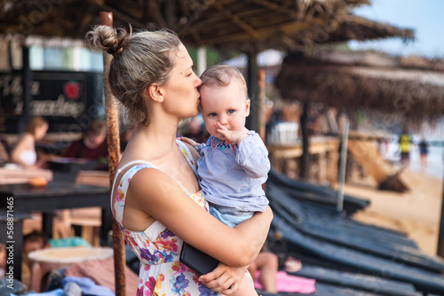 Happy mom kissing baby in arms on recreation at tropical beach background. Female with little child in summer travel vacation. Concept of motherhood and enjoying with children. Copy text space for ad