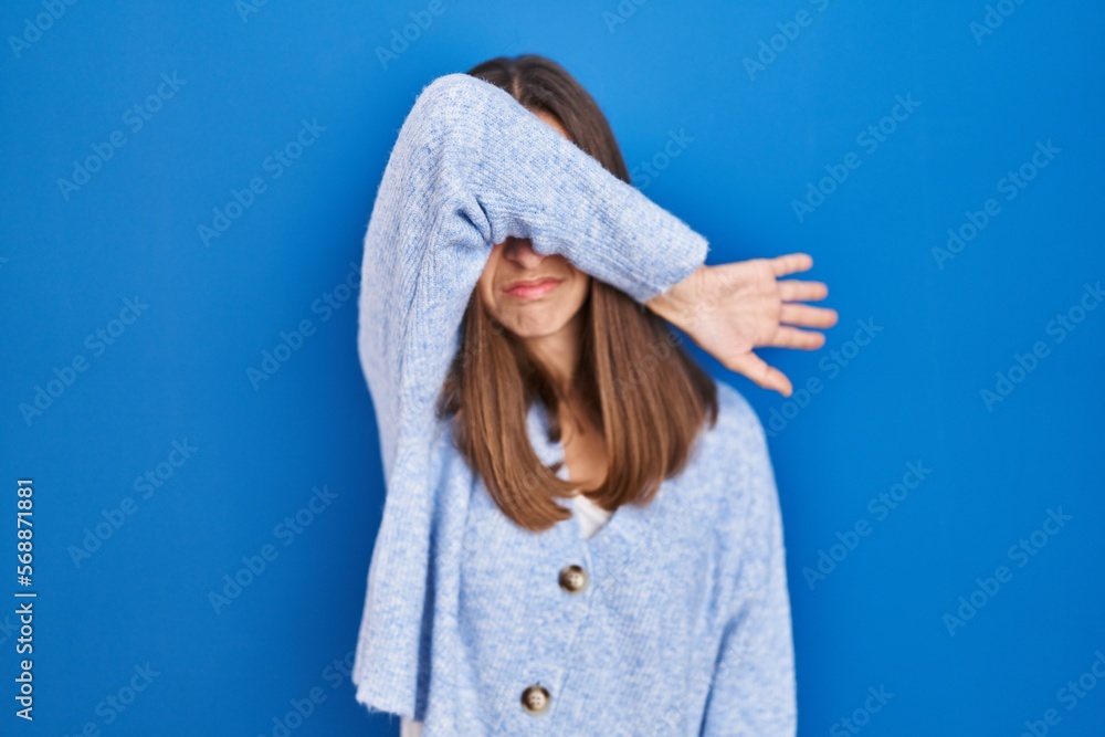 Obraz premium Young woman standing over blue background covering eyes with arm, looking serious and sad. sightless, hiding and rejection concept