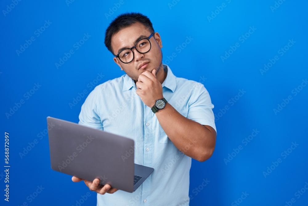 Chinese young man using computer laptop with hand on chin thinking about question, pensive expression. smiling with thoughtful face. doubt concept. Chinese young man using computer laptop with hand on chin thinking about question, pensive expression. smiling with thoughtful face. doubt concept.