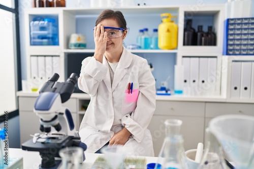 Hispanic girl with down syndrome working at scientist laboratory covering one eye with hand, confident smile on face and surprise emotion.