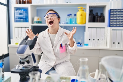 Hispanic girl with down syndrome working at scientist laboratory crazy and mad shouting and yelling with aggressive expression and arms raised. frustration concept.