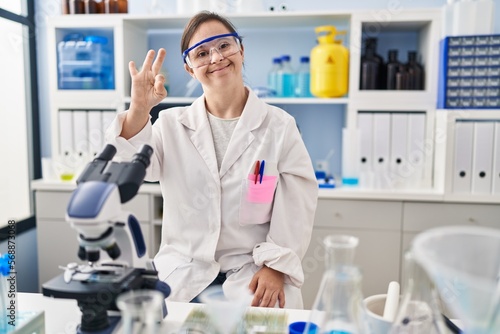 Hispanic girl with down syndrome working at scientist laboratory showing and pointing up with fingers number three while smiling confident and happy.