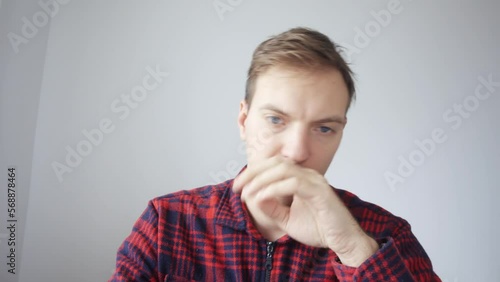 A man in a red shirt is sitting in front of a webcam. He picks his nose during a video conference. He gets scared, pulls his hand away, apologizes. View from the laptop webcam.
