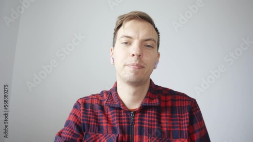 A young man finishes an online meeting, puts down his wireless headphones and leaves. A man in a red shirt communicates via laptop video communication. View from the laptop webcam.