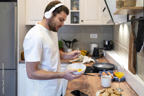 Man in white tee shirt and headphones standing in kitchen cooking omelet for breakfast and listening music.